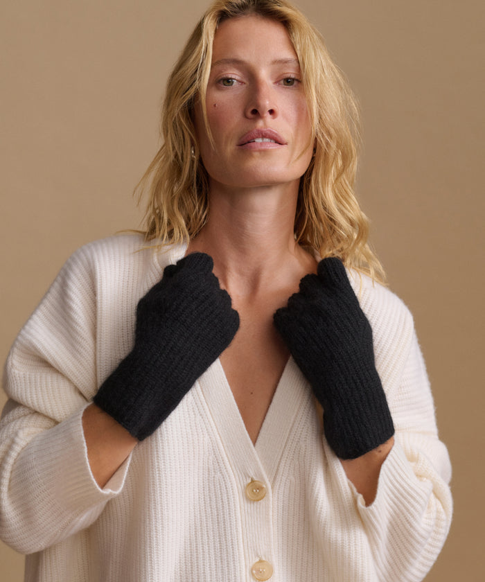 A woman wearing black knit fingerless gloves over a cream ribbed cardigan, posed against a solid beige background.
