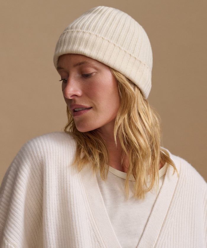 A woman wearing a cream ribbed knit beanie and a matching cream knit cardigan, shown in a close-up portrait against a warm beige background.