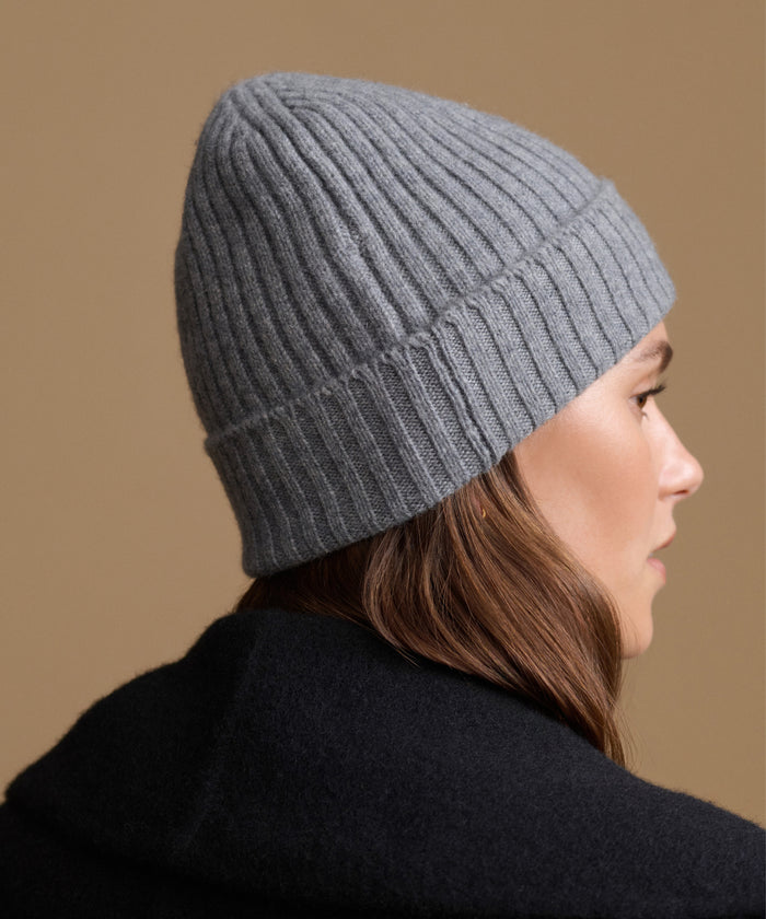 A woman shown in profile wearing a ribbed knit beanie hat in heather gray, paired with a dark coat, against a solid warm tan background.