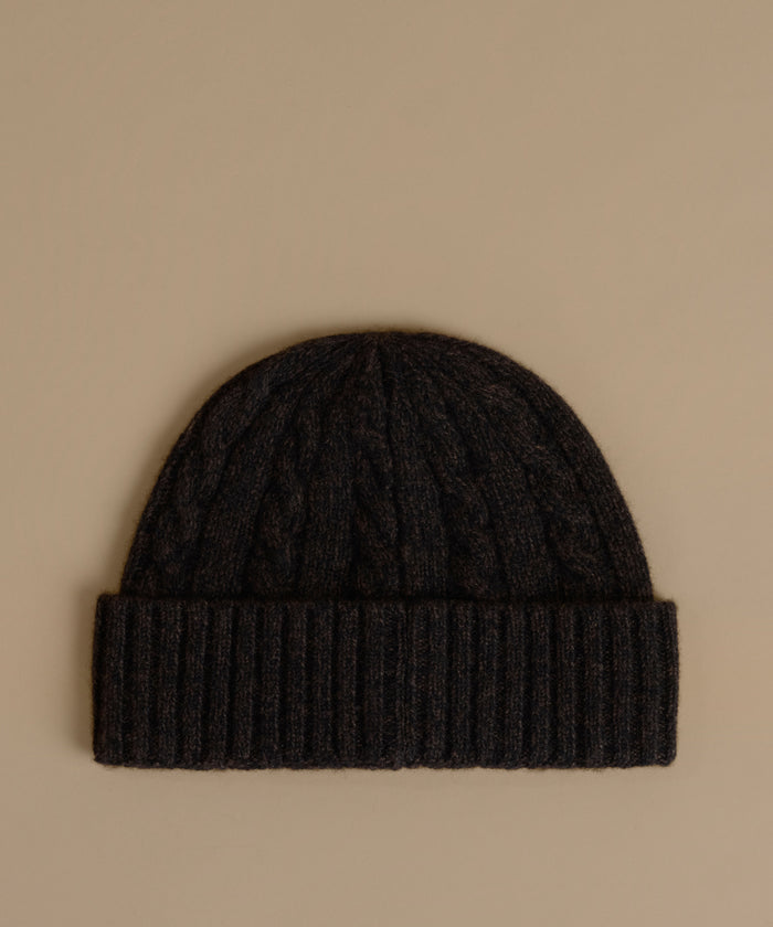A dark charcoal cable-knit beanie with a ribbed cuff, displayed flat against a solid beige background.