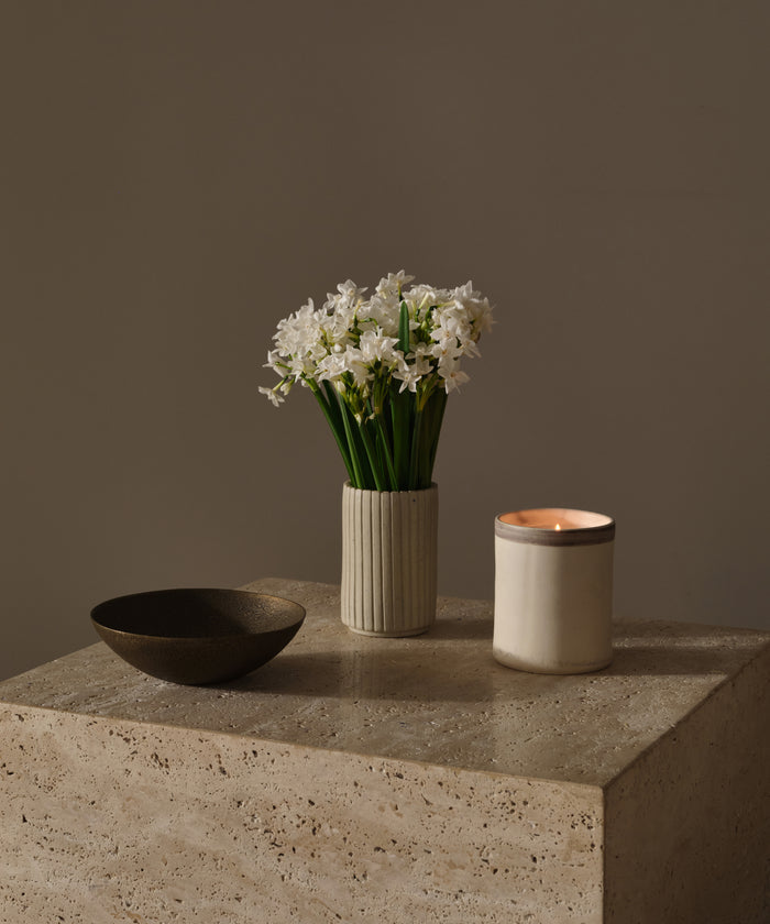 A ceramic vase with white flowers, a lit candle in a cream holder, and a Cast Iron Bowl are displayed on a stone surface against a neutral background.
