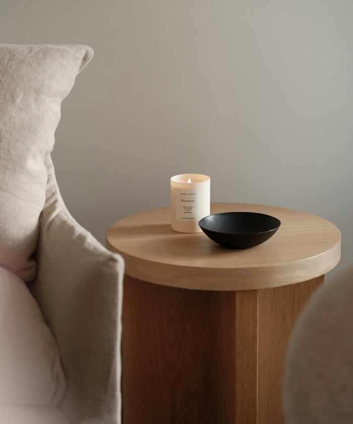 A round wooden side table with a lit white candle and a Cast Iron Bowl sits beside a beige cushioned chair against a light gray wall.