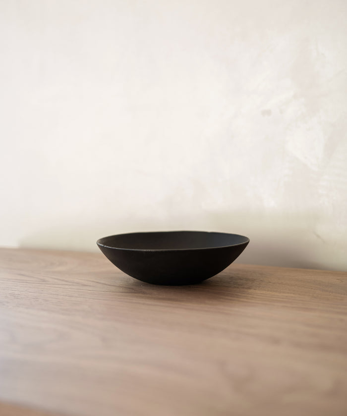 A minimal scene features the Cast Iron Bowl by Blackcreek Mercantile, crafted in the USA. The simple round bowl sits on a light wood surface against an off-white wall, highlighted by soft lighting.