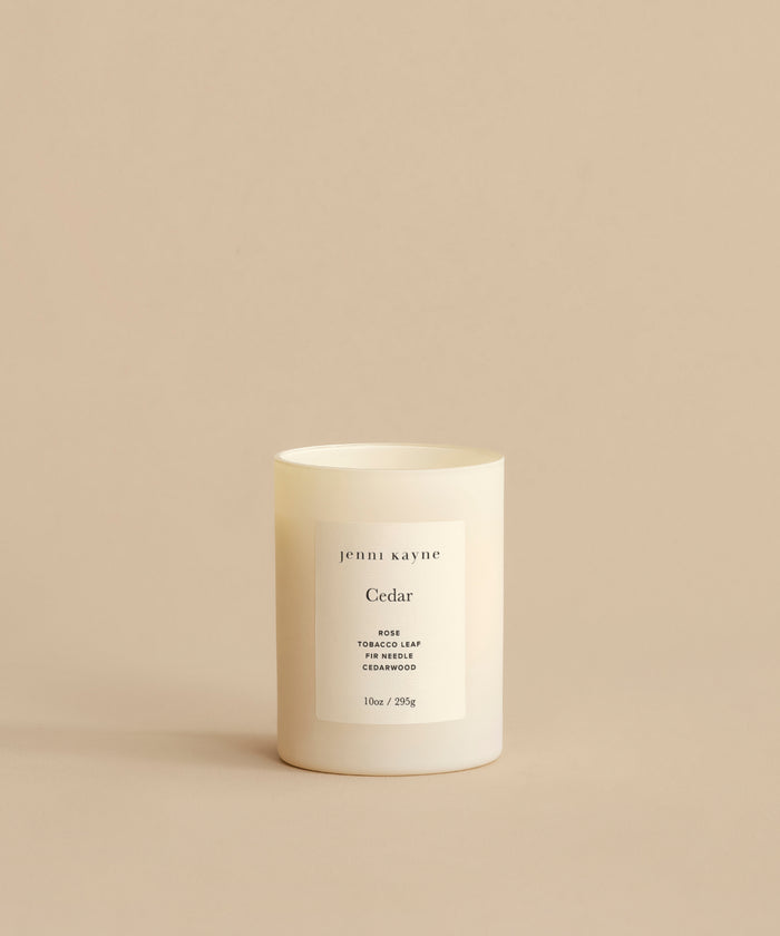 The Cedar Glass Candle, hand-poured in Los Angeles, features a minimalist white design and scent notes of rose, tobacco leaf, and cedarwood. It is shown on a beige surface with a matching beige background.