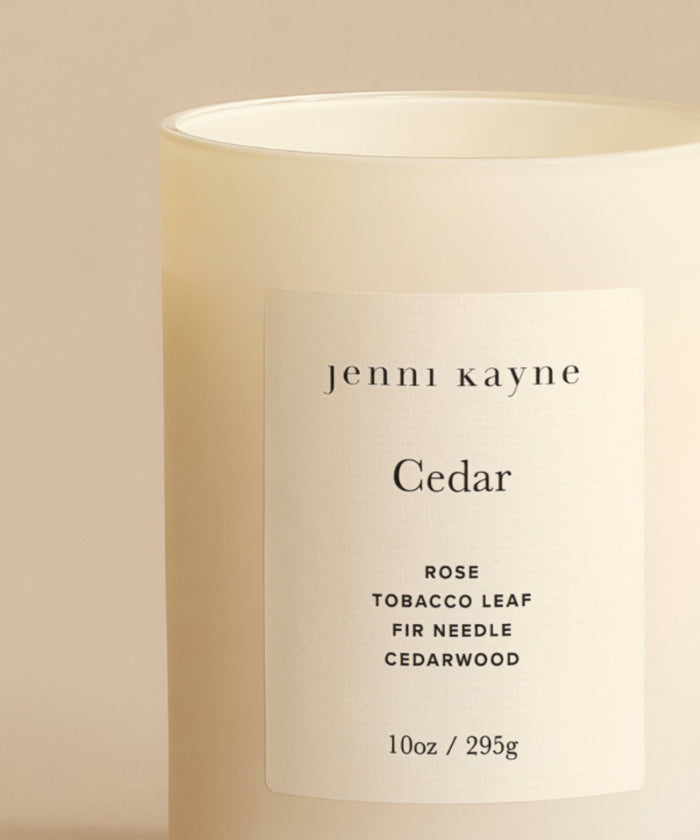 The Cedar Glass Candle from Jenni Kayne, a 10oz cream-colored candle hand-poured in Los Angeles, features rose, tobacco leaf, fir needle, and cedarwood notes. One of Oprah’s Favorite Things, it’s pictured against a neutral background.