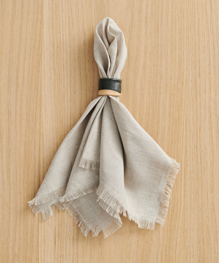A beige fabric napkin with frayed edges is gathered through a Ceramic Napkin Rings Set of 4, handmade with a black glaze, and placed on a light wood surface.
