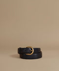 A black leather belt with a polished gold-tone metal buckle, neatly coiled on a warm beige studio background.