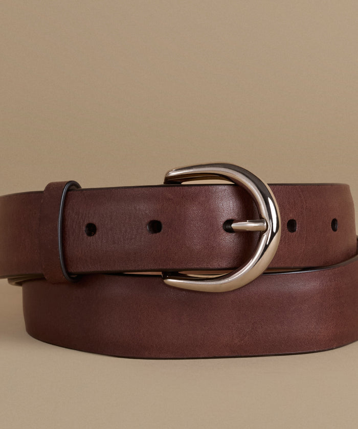 A dark brown leather belt with a polished silver-tone metal buckle, neatly coiled on a solid beige background.