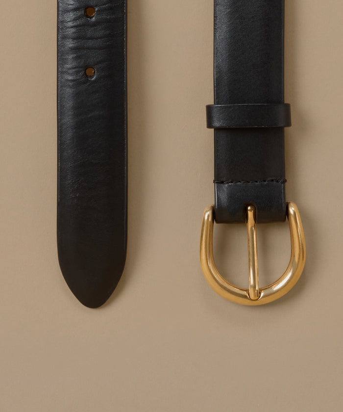 A close-up of a black leather belt laid flat on a warm beige background, showing the textured strap, adjustment holes, and a polished gold-tone metal buckle.