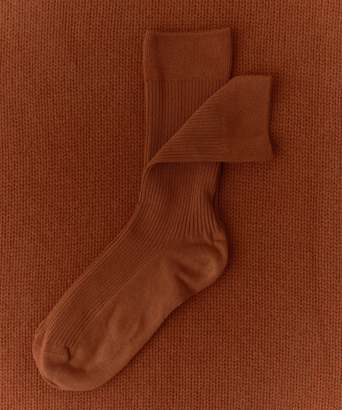 A pair of Classic Sock in rust, made from a lightweight, breathable lyocell blend, laid on a matching rust knitted fabric background with one sock partially overlapping the other.