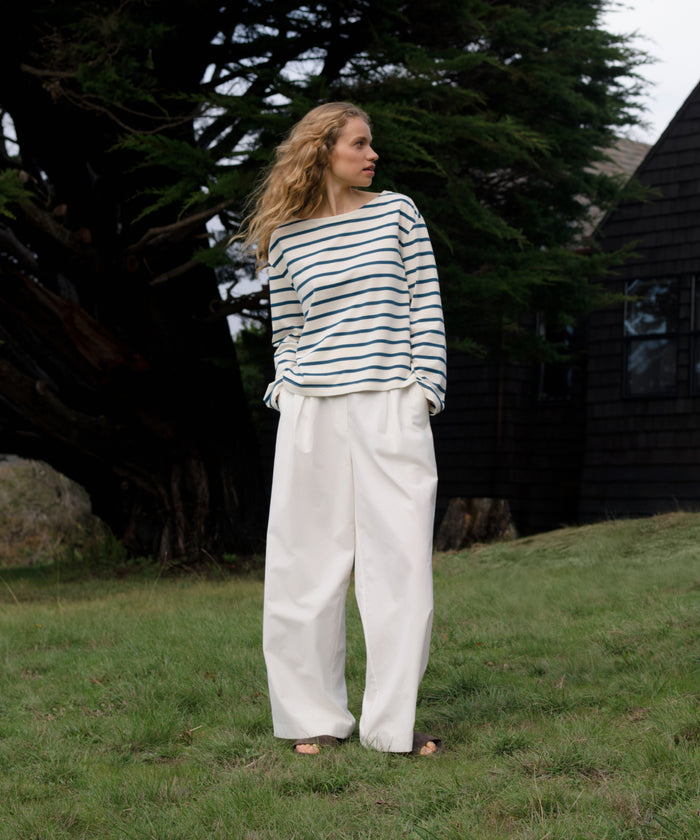 Woman wearing a striped shirt and white pants standing in a grassy area with trees and a building in the background.