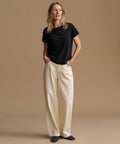 A woman stands against a plain beige studio background wearing a black short-sleeve crew-neck T-shirt and cream wide-leg trousers, with her hands in the trouser pockets.
