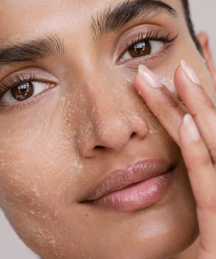 A close-up of someone gently cleansing their face with the Cleanse and Polish Duo, visible exfoliant particles enhancing their smooth, clear skin. Well-groomed brows and natural makeup complete their fresh, radiant look.