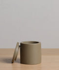 The Coast Canister, a cylindrical matte taupe handcasted resin container with matching lid, rests on light wood in front of a plain light grey background as part of the Coast Collection.