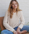 A woman with blonde hair sits barefoot on a wooden chair, wearing a cream-colored ribbed knit sweater and blue jeans against a white brick wall.