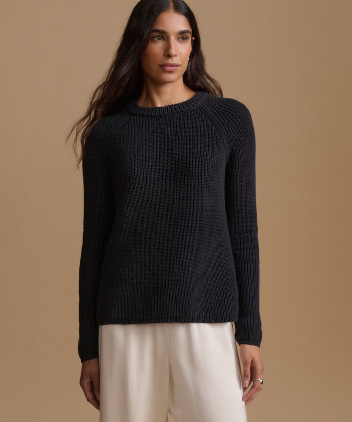 A woman wearing a black knit sweater and cream trousers, standing against a solid beige studio background.