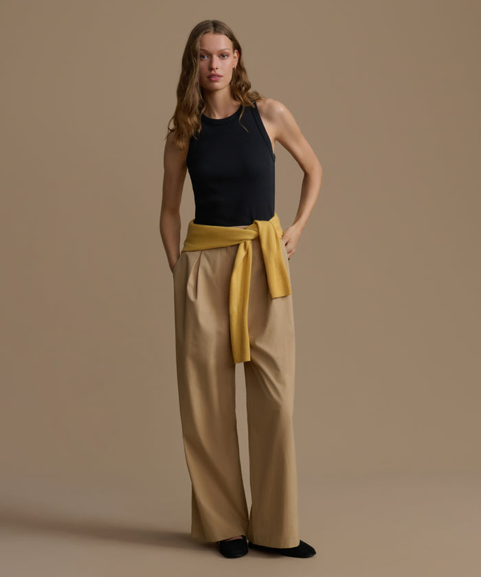 Woman wearing a black top and beige pants with a yellow belt against a brown background