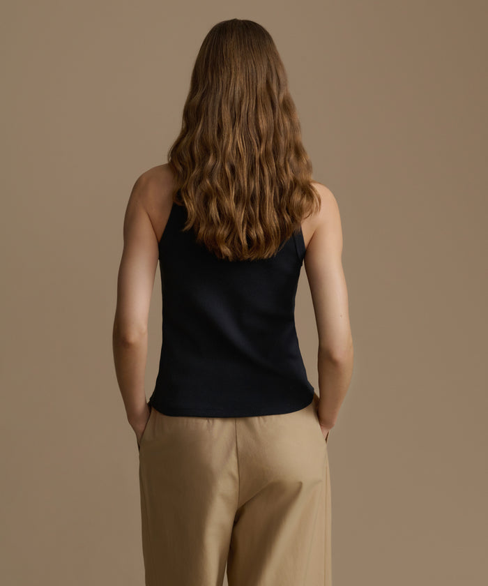 Person wearing a black sleeveless top and beige pants against a brown background