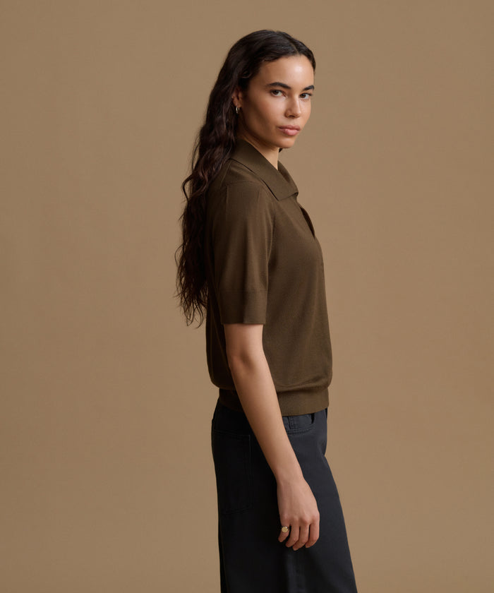 A woman in side profile wearing a short-sleeve knit polo shirt in dark brown and dark navy pants, set against a solid warm beige studio background.