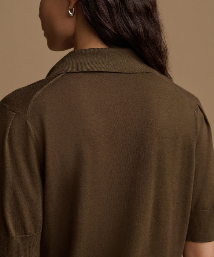 Close-up back view of a woman wearing a short-sleeve knit top in a dark olive brown color, set against a solid warm beige background.
