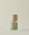 Against a beige backdrop, the clear glass Cuir Rose Eau De Parfum bottle—with a light wood-grain cap and green label—showcases its clean fragrance, blending floral and woody notes.