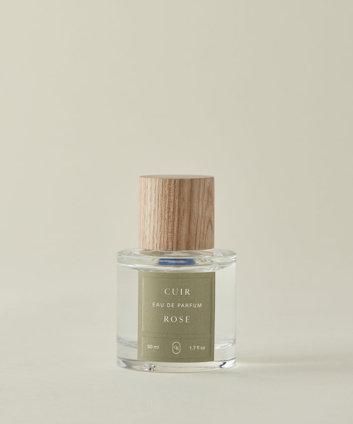 Against a beige backdrop, the clear glass Cuir Rose Eau De Parfum bottle—with a light wood-grain cap and green label—showcases its clean fragrance, blending floral and woody notes.