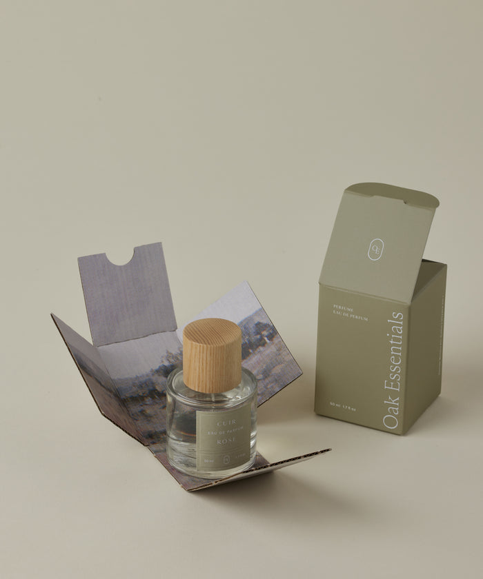 A glass bottle of Cuir Rose Eau De Parfum with a wooden cap rests in an open box with scenic artwork, beside a closed olive-green Oak Essentials box on a beige background, evoking the floral and woody notes of this clean fragrance.