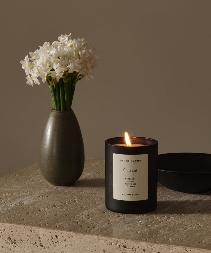 A small brown vase with white flowers and a lit Currant Glass Candle, made from natural coconut soy wax, rest on a stone surface against a neutral background.