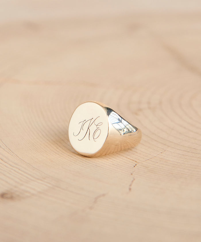 A gold signet ring with engraved initials JKC in elegant script rests on a light wooden surface—a timeless piece, expertly customized with our Custom Engraving Service.