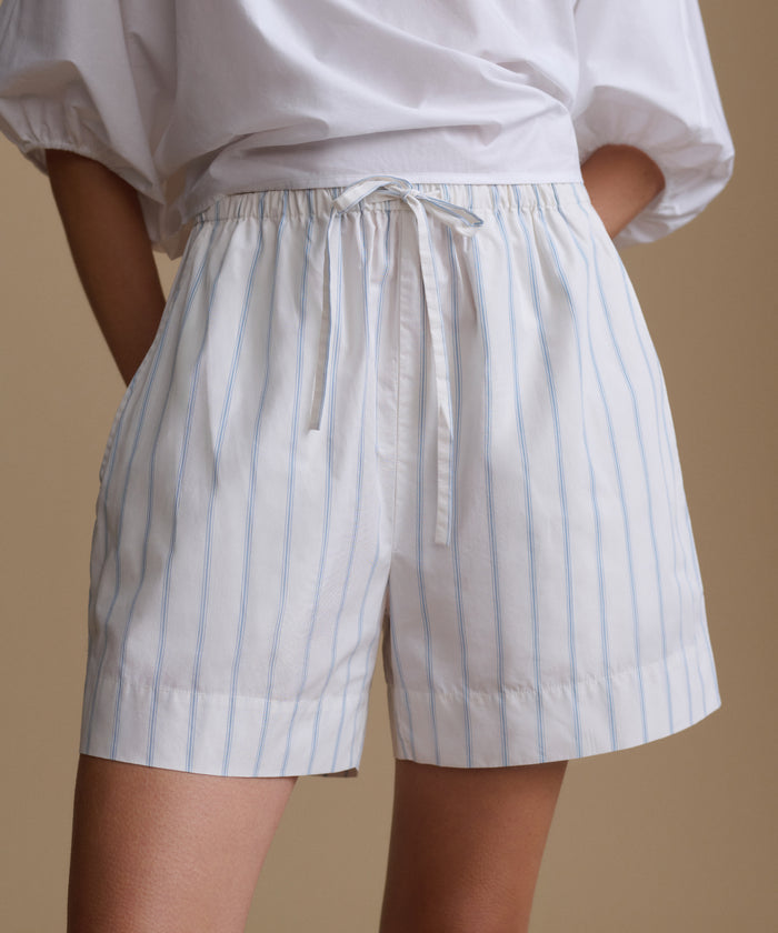 Woman wearing a white blouse with white and blue stripe shorts against a beige background.