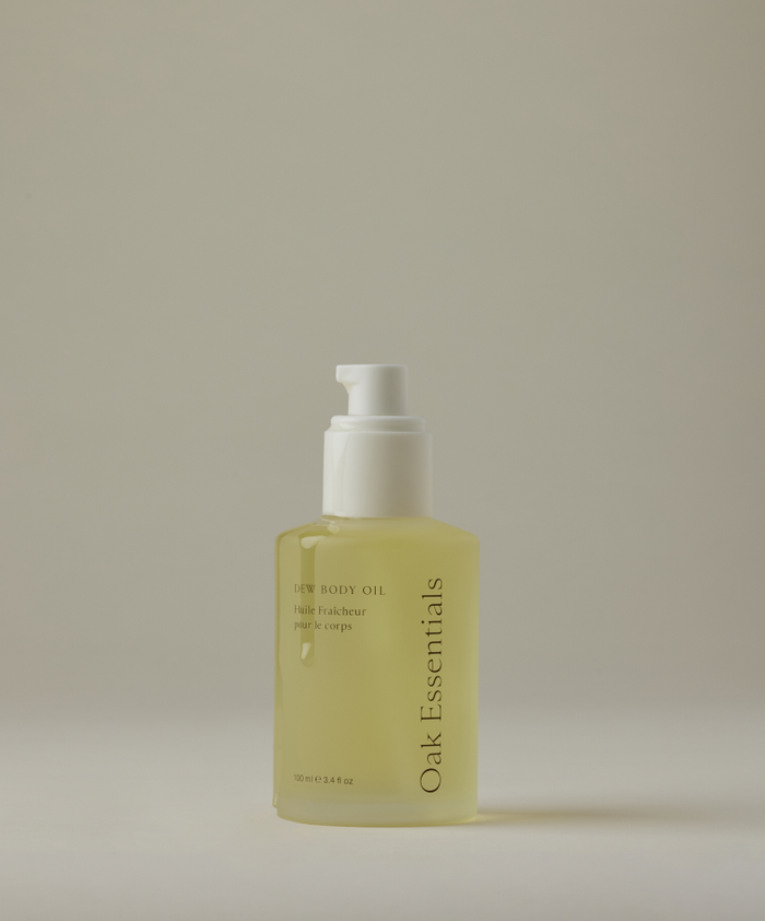 A clear pump bottle of Dew Body Oil, filled with yellow moisturizing oil, stands upright against a plain, light background.