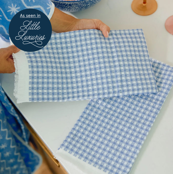 A person holds a folded blue and white gingham check fabric napkin with white fringe, alongside another matching napkin on a white table. A dark blue circular badge in the top left reads 'As seen in Little Luxuries'.