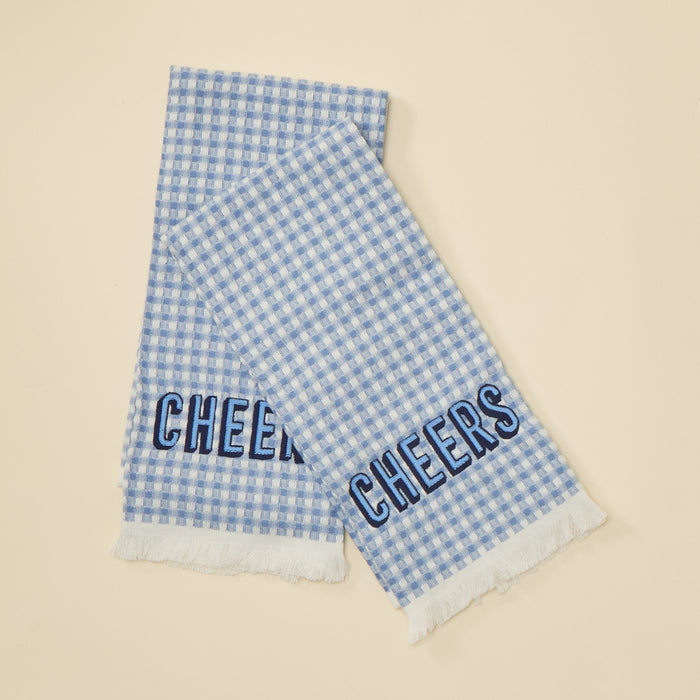 A light blue and white gingham checkered scarf with white fringe, featuring the word 'CHEERS' embroidered in navy blue outline lettering on one end, displayed on a solid beige background.