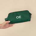 A hand holds a sleek Hunter Green Dopp Kit featuring a personalized "OE" monogram in crisp, white embroidered letters.