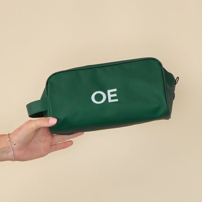 A hand holds a sleek Hunter Green Dopp Kit featuring a personalized "OE" monogram in crisp, white embroidered letters.