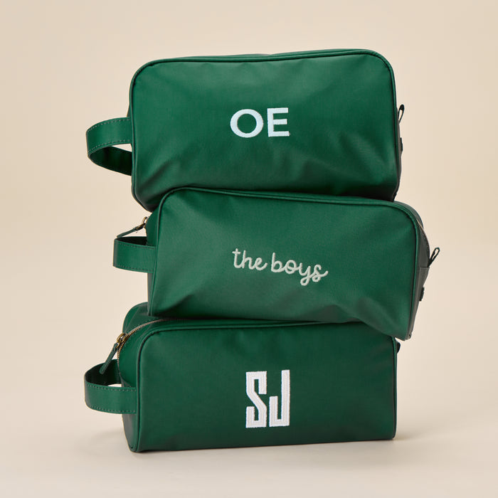 A hand holds a sleek Hunter Green Dopp Kit featuring a personalized "OE" monogram in crisp, white embroidered letters.