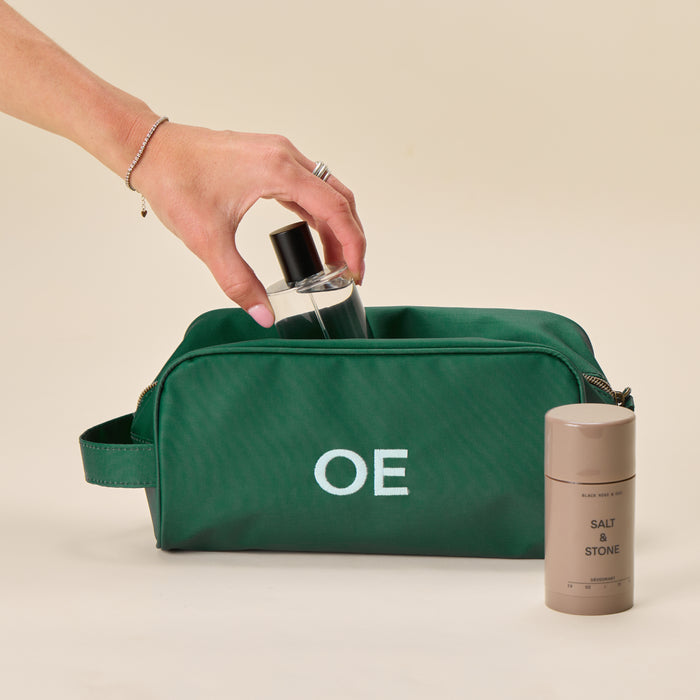 A hand places a bottle into a Hunter Green Dopp Kit with a white "OE" monogram, next to a Salt & Stone deodorant stick.
