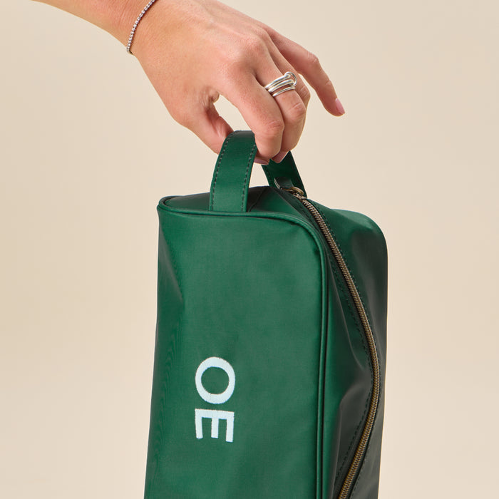A hand holding a structured Hunter Green Dopp Kit featuring a white "OE" monogram, shown against a neutral background.