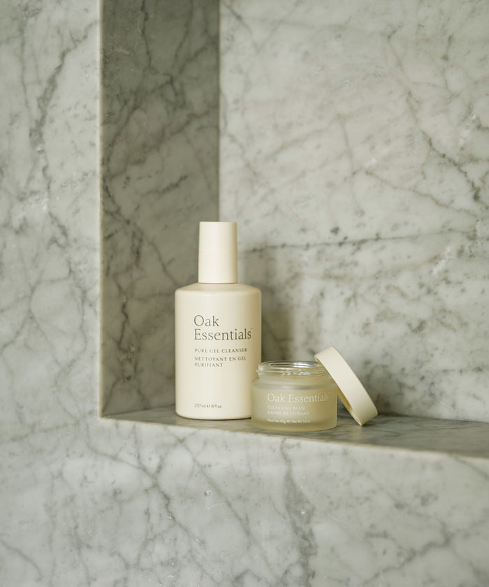 The Double Cleanse Duo by Oak Essentials, featuring minimalistic, soft beige packaging, is displayed on a marble shelf with a gray veined background, exuding effortless elegance.