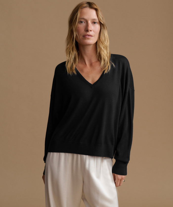 A woman modeling a black long-sleeve V-neck sweater paired with cream-colored pants, set against a solid warm beige studio background.