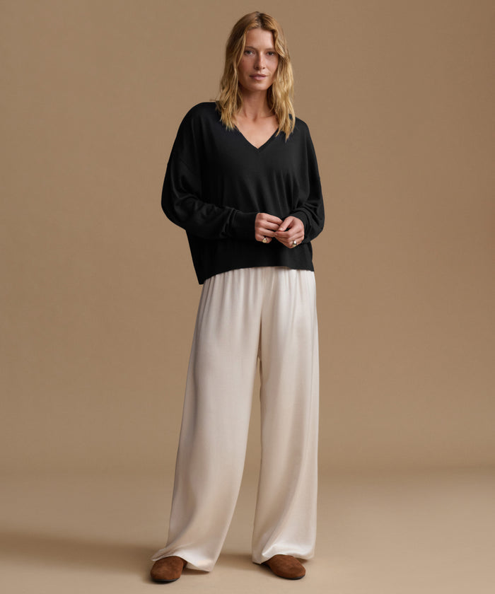 A woman modeling a black long-sleeve V-neck top and cream wide-leg trousers against a solid beige studio background.