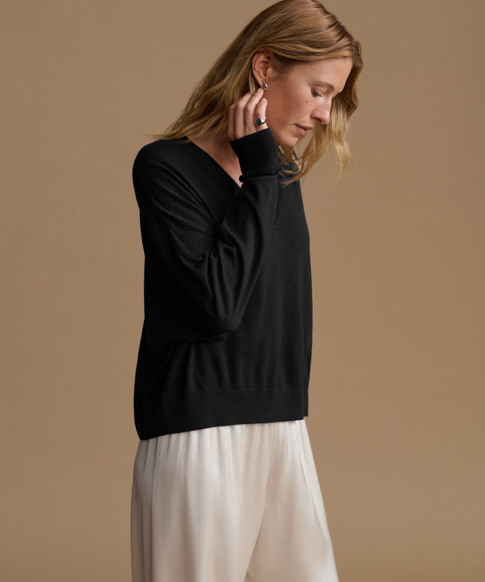 A woman wearing a black long-sleeve knit sweater and white wide-leg trousers, shown from the waist up against a solid warm beige studio background.