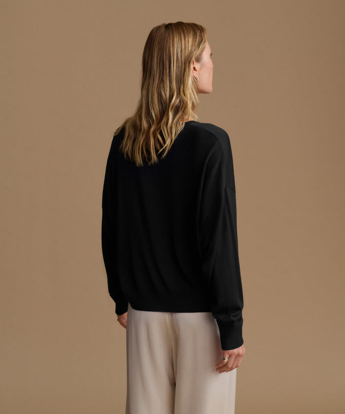 A woman with blonde hair is shown from the back wearing a black long-sleeve knit top and light beige wide-leg trousers, set against a solid warm beige studio background.