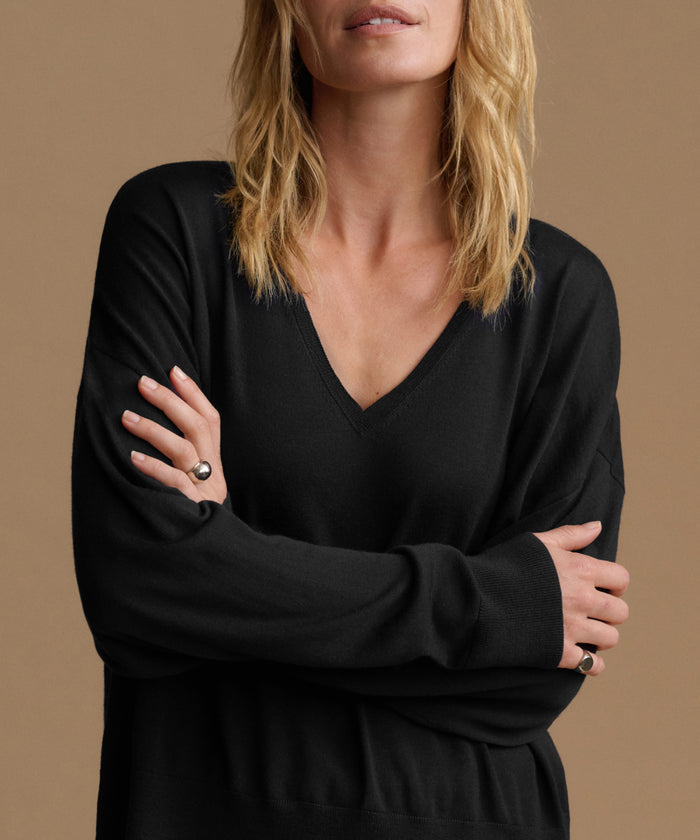 A woman wearing a black long-sleeve V-neck knit top, shown from the lips down with her arms crossed. She is wearing silver rings on her fingers.