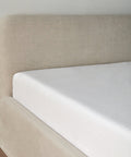 A close-up of a neatly made bed with a light beige upholstered headboard, showcasing the Dusk Linen Fitted Sheet—lightweight, comfortable, and made in Portugal—set against a plain light-colored wall.