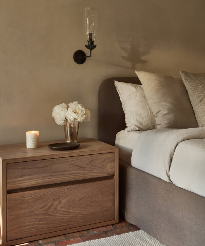 A cozy bedroom scene with a wooden nightstand, candle, white flowers, metal tray, and a bed dressed in the Dusk Linen Fitted Sheet made in Portugal, with a wall-mounted sconce overhead.