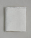 The Dusk Linen Flat Sheet, in crisp white, lies neatly folded on a light gray surface. Its soft, smooth fabric and minimal texture offer understated elegance—ideal for those who love luxurious bedding.