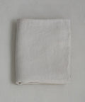 The Dusk Linen Flat Sheet, in light beige natural linen, is neatly folded and lies flat on a textured pale surface, highlighting the understated elegance of luxury bedding.