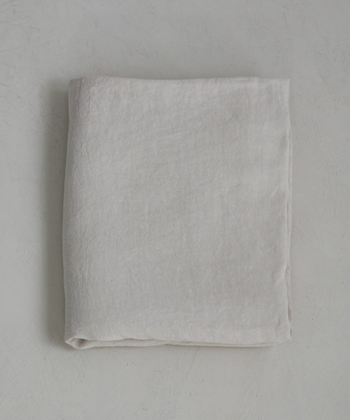 The Dusk Linen Flat Sheet, in light beige natural linen, is neatly folded and lies flat on a textured pale surface, highlighting the understated elegance of luxury bedding.