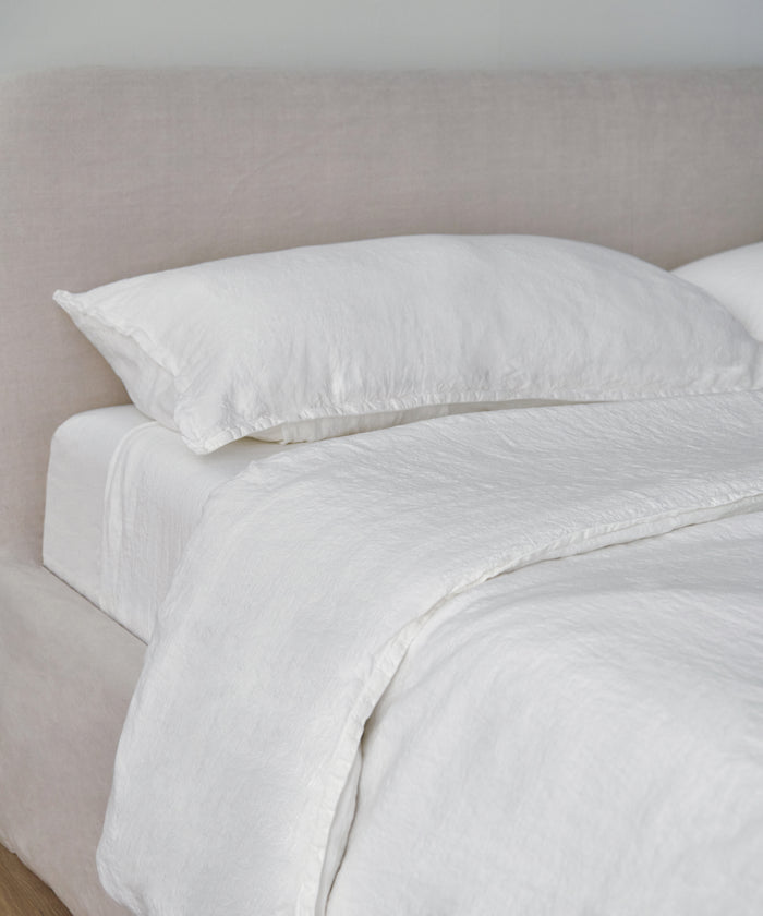 A neatly made bed with a light beige upholstered headboard, dressed in luxury bedding including the Dusk Linen Flat Sheet, pillow, and duvet, creates a clean and minimalistic look.